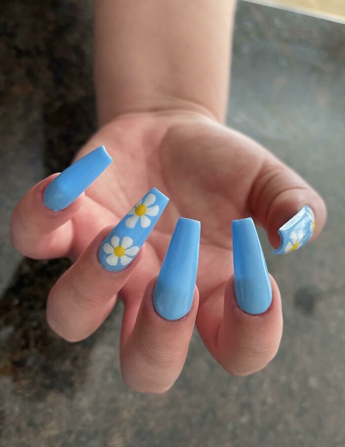 Close-up of long blue nails with floral designs showcasing creative nail art ideas on a hand against a dark background.