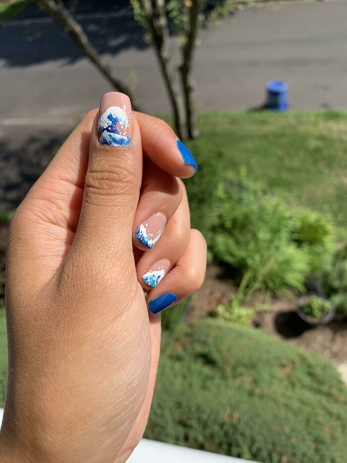 Hand with nail art ideas featuring blue and white wave designs and solid blue polish outdoors with blurred greenery background.