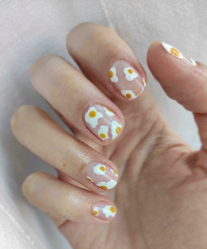 Close-up of hand with clear nails featuring white and yellow floral nail art ideas on a soft fabric background.