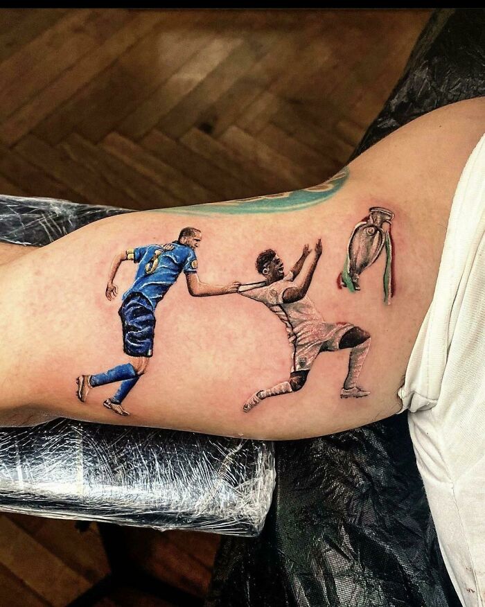 Quality tattoo of soccer players with poor design on arm, featuring two athletes and a trophy.
