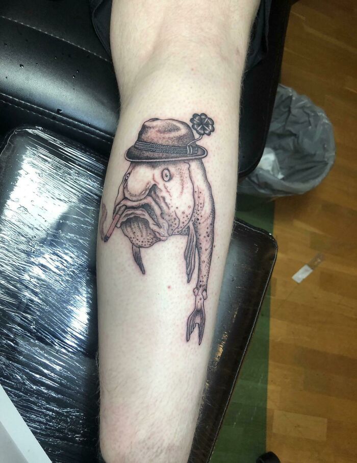 Tattoo design of a fish wearing a hat, poorly executed on a person's leg.