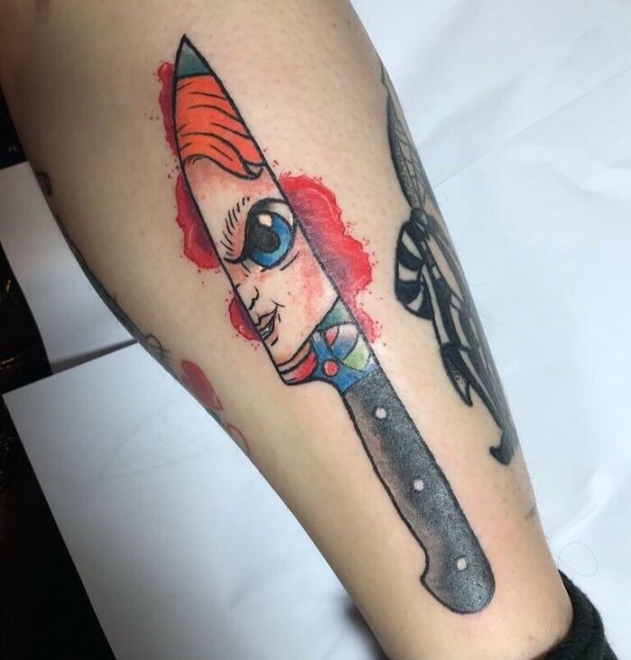 Trippy tattoo of Chucky's face on a knife with vibrant colors and bold design.