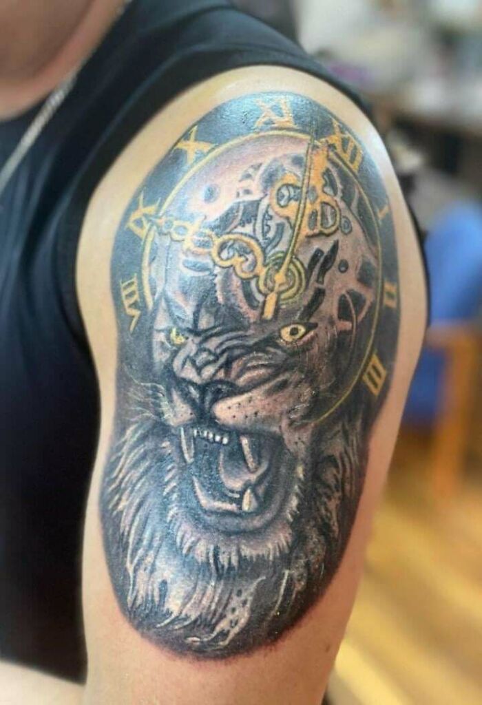 Tattoo of a lion with a clock on a person's upper arm, showcasing bold colors and intricate details.