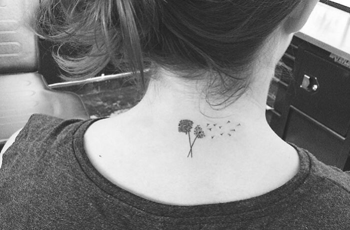 Minimal tattoo design of dandelions on the nape of a woman's neck in a monochrome setting.