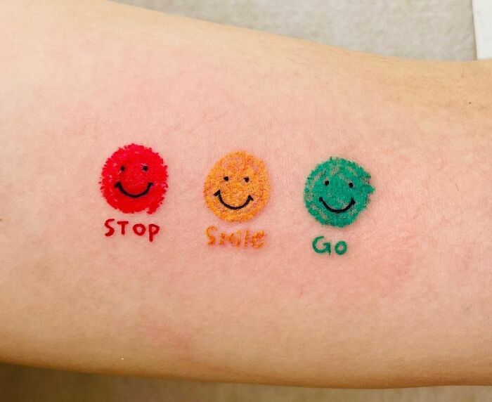 Minimal tattoo of three smiley faces in red, orange, and green with text "Stop", "Smile", "Go" on skin.