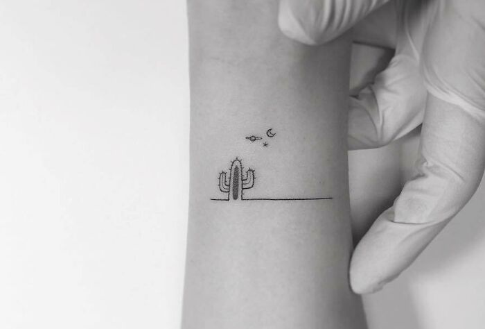 Minimal tattoo design of a cactus under stars and a crescent moon on an arm, held by a gloved hand.