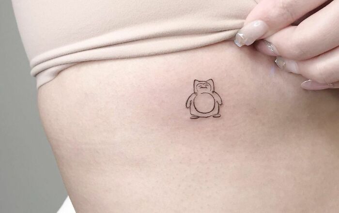 Minimal tattoo design of a cartoon character on a person's side, with a hand gently touching the skin.