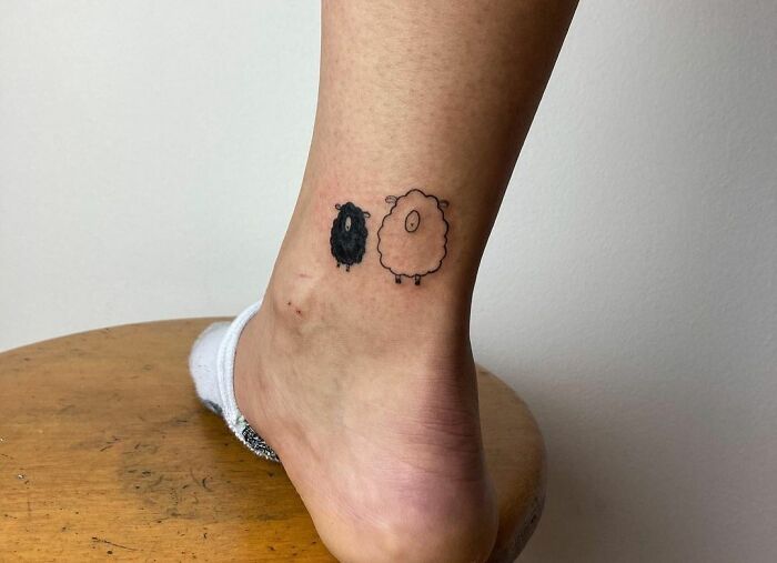 Minimal tattoo of two cartoon sheep on an ankle, one black and one white, showcasing creative simplicity.