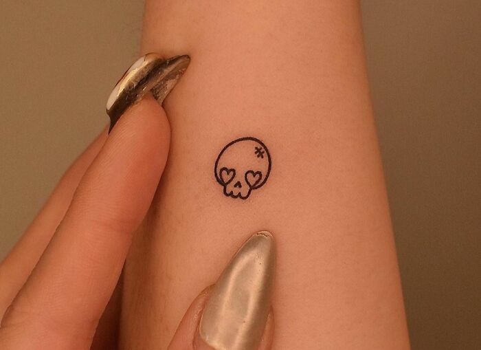 Minimal tattoo design of a small skull with heart eyes on an arm.