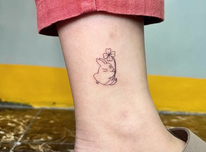 Minimal tattoo design of a small rabbit holding a flower on an ankle.