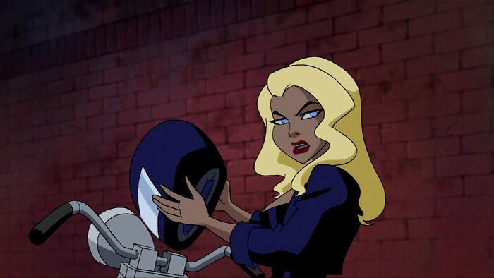 Blonde female cartoon character with helmet, showcasing fame and strength, standing by a motorcycle against a brick wall.