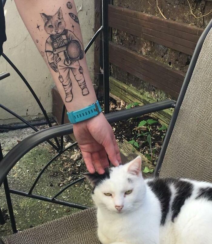 Tattoo of an astronaut cat on a person's arm, with a white and black cat being petted, symbolizing adorable pet tattoos.