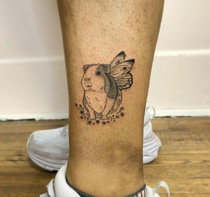 Guinea pig tattoo with butterfly wings on leg, showcasing adorable pet art.