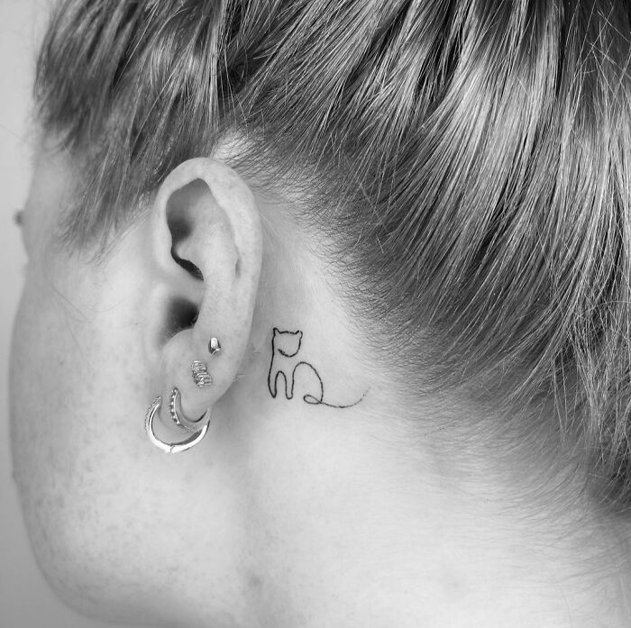 Fine-line tattoo of a cat behind the ear with earrings and short hair.