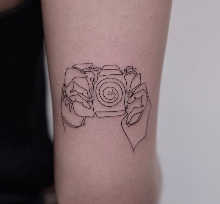 Fine-line single-line tattoo of hands holding a camera on an arm.