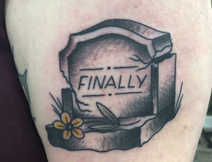 Funny leg tattoo of a c*****d grave with "FINALLY" inscription and small flower.