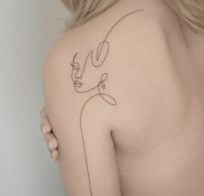 Fine-line tattoo of a face on a person's shoulder, showcasing single-line artistry.