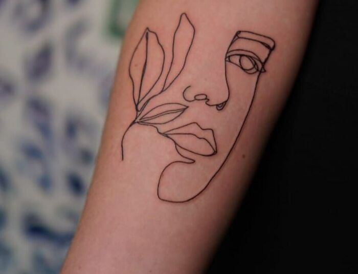 Fine-line tattoo of an abstract face with leaves on an arm.