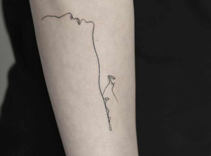 Single-line tattoo on forearm depicting a face in fine-line perfection on a black background.