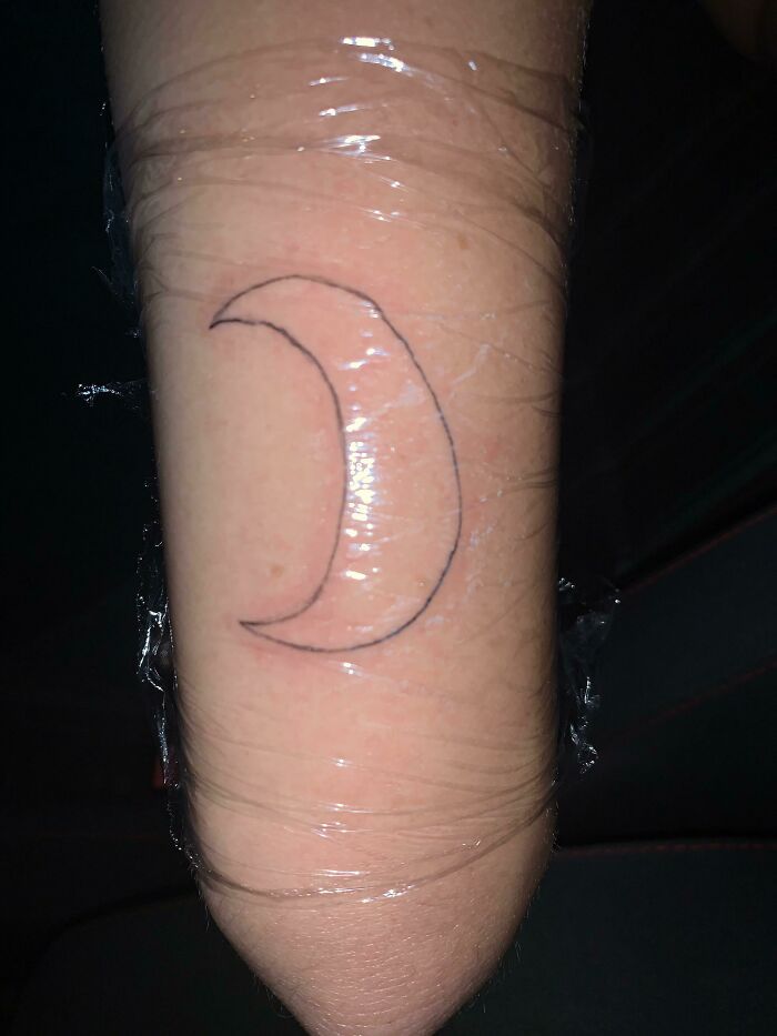 Minimalist crescent moon tattoo, humorously resembling a banana shape, on a forearm wrapped in protective plastic.