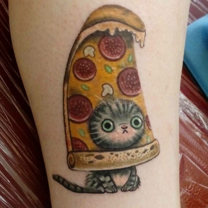 Funny tattoo of a cat under a slice of pizza with wide eyes on the arm.