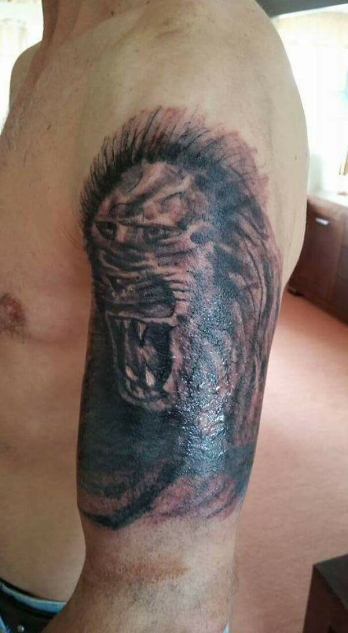 Funny tattoo of a lion's face on an upper arm, featuring exaggerated expression and bold outlines.