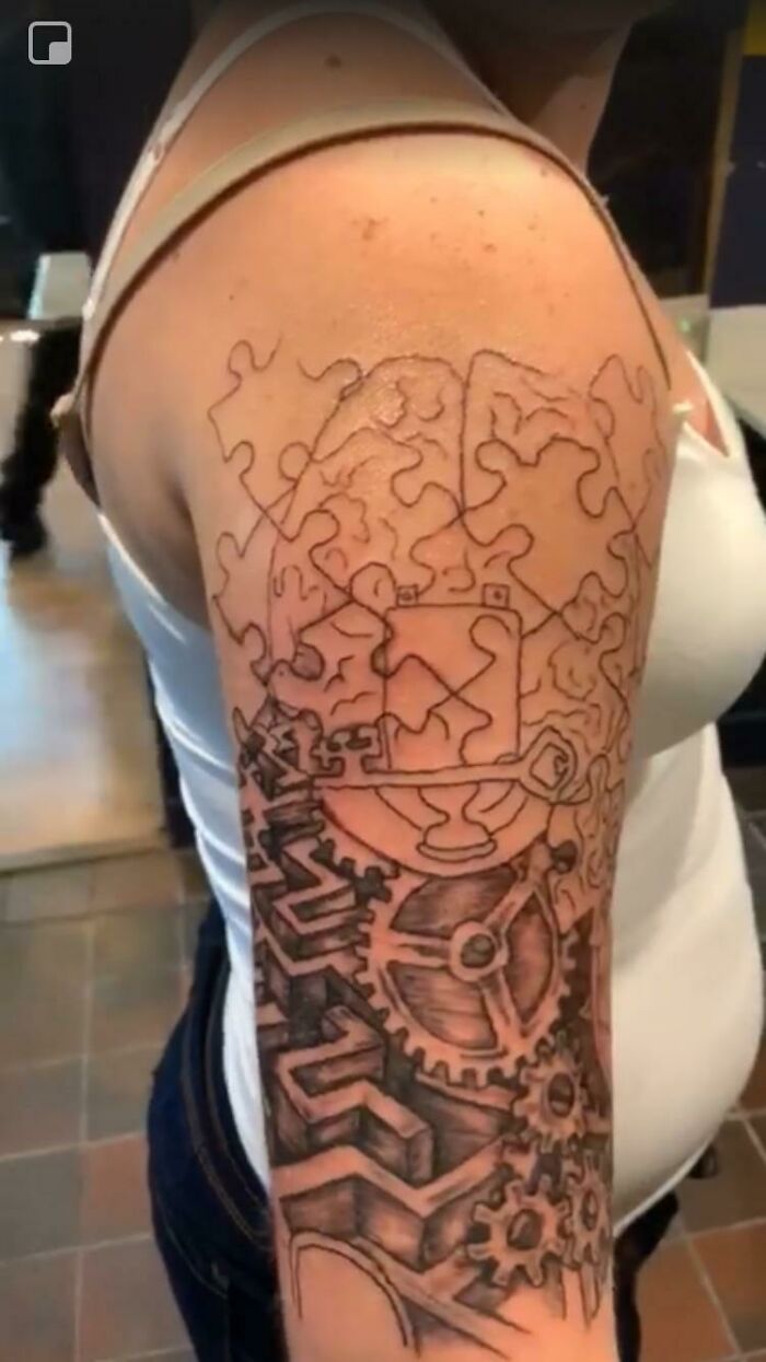 Puzzle piece and gear-themed tattoo on upper arm, featuring intricate black ink design elements.