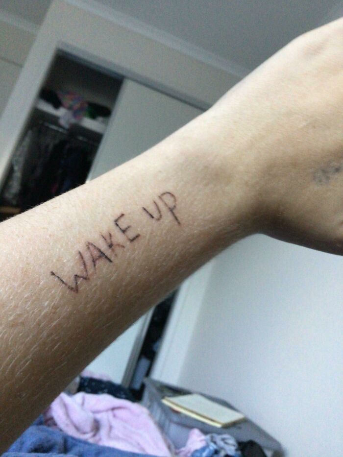 Funny tattoo on forearm with the phrase "Wake Up" in a casual room setting.