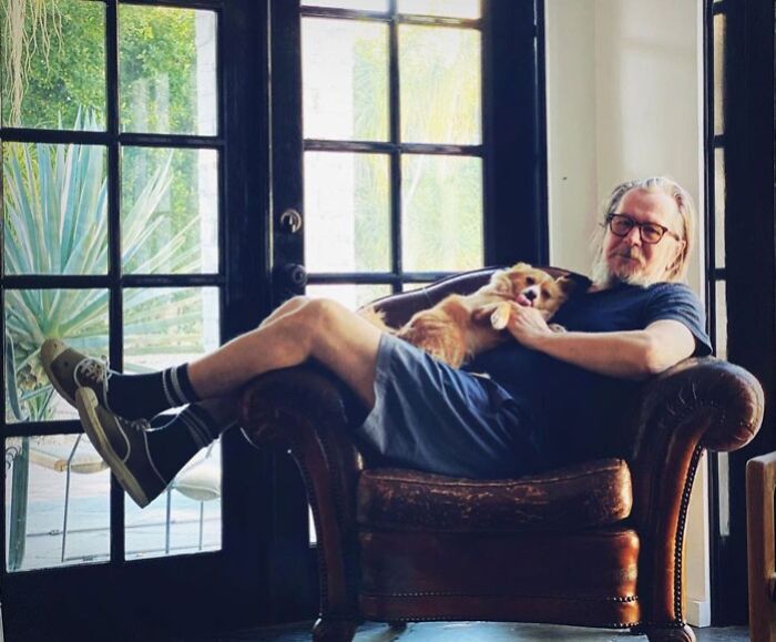 Gary Oldman sitting in a chair holding his dog, relaxing indoors, representing famous people married multiple times.