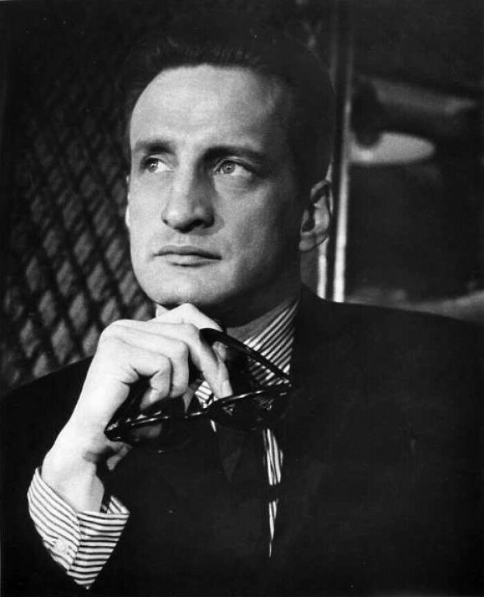 George C. Scott wearing a suit, holding glasses thoughtfully, representing famous people who got married multiple times.