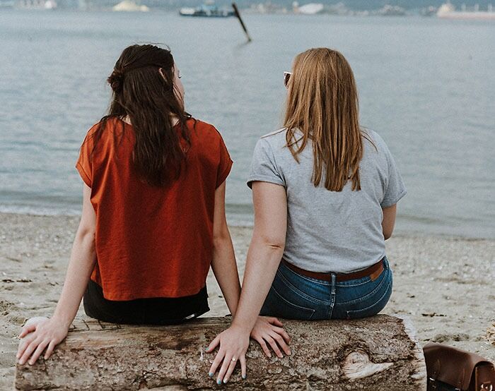 Two women sitting on a log by the beach holding hands, enjoying a relaxed moment perfect for texting games.