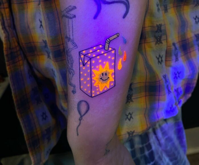 UV-reactive tattoo of a juice box with a smiling sun, vibrant colors, and drip details on arm under UV light. Trippy tattoos theme.