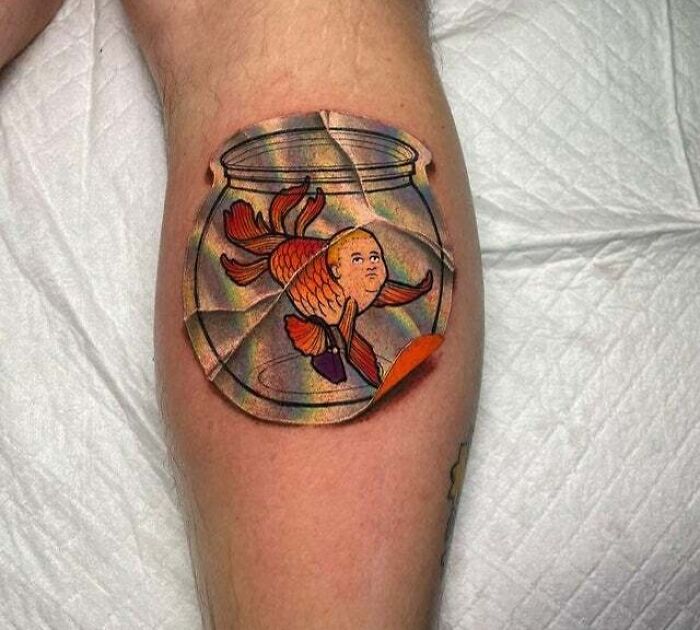 Trippy tattoo of a goldfish with a human face inside a c*****d fishbowl on a person's leg.