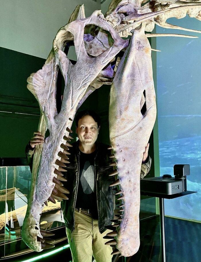 Man posing inside large prehistoric reptile skull jaws, showcasing nature was metal with intense fossil display.