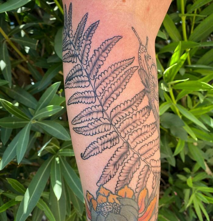 Fern tattoo design symbolizing mother-child bond, inked on forearm, shown against green leaves.