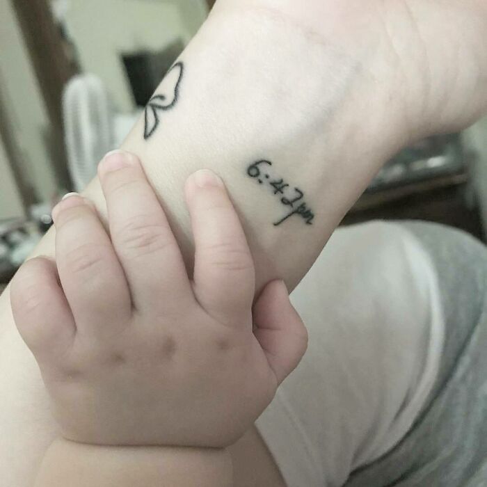 Baby's hand touching a wrist with a mother-child bond tattoo, featuring a time and butterfly outline.