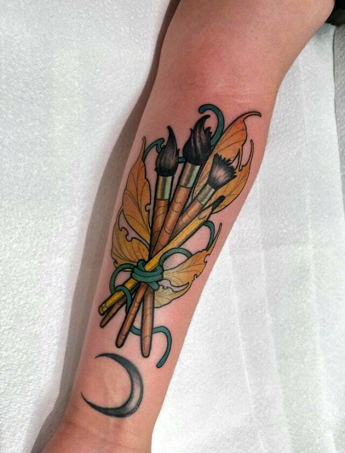Tattoo of paintbrushes and leaves with a crescent moon, symbolizing the mother-child bond.