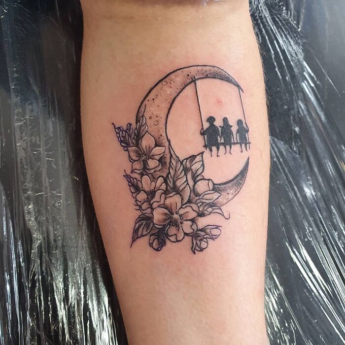 Mom tattoo featuring a crescent moon with flowers and silhouettes of children on a swing, symbolizing the mother-child bond.