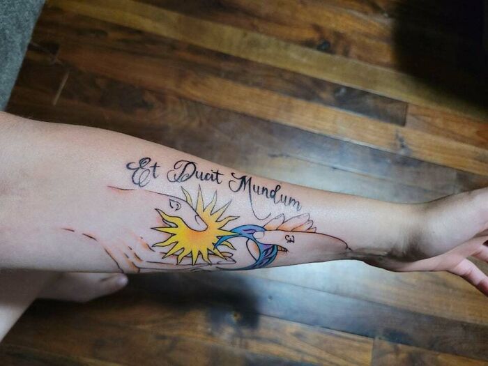 Forearm tattoo with vibrant sun and moon, symbolizing the mother-child bond, and the phrase "Et Ducit Mundum."