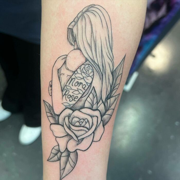 Mother-child bond tattoo with a woman holding a baby, roses, and the names "Nora Rose" inscribed.