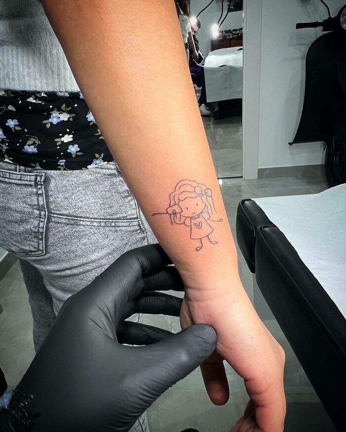 Mom tattoo on forearm featuring a child’s drawing of a girl, symbolizing the mother-child bond.