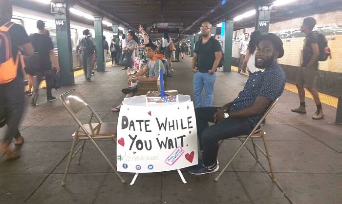 Man sitting at a table with a sign offering to date while waiting in a busy New York City subway station.