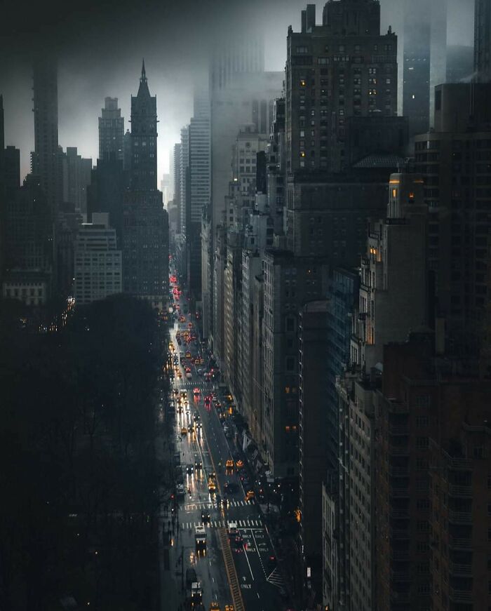 Moody evening view of New York City street lined with tall buildings and glowing car lights in rain.