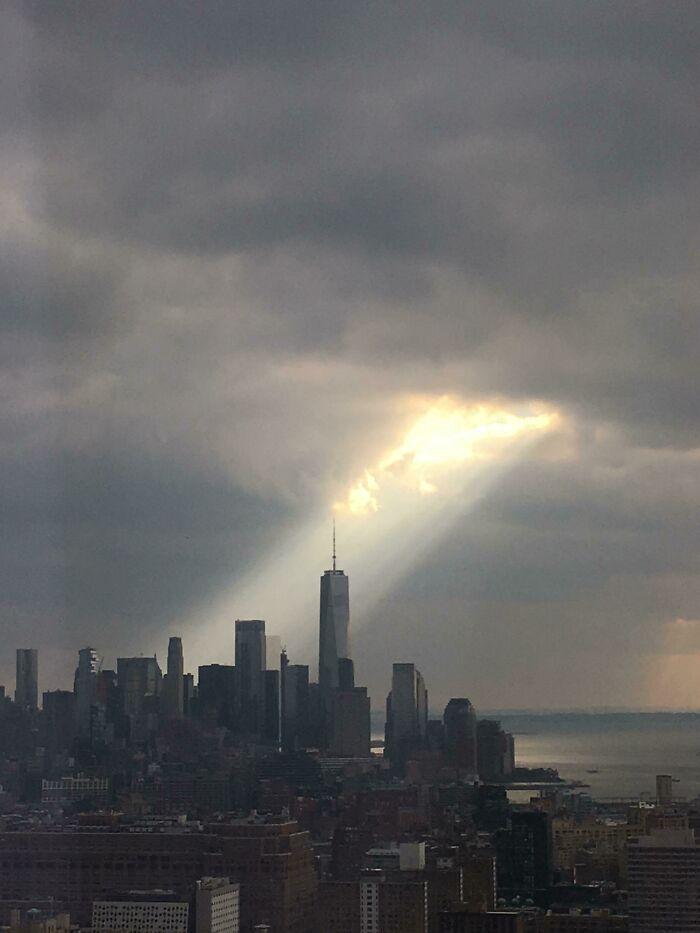 Dark clouds over New York skyline with a striking sunbeam illuminating One World Trade Center uniquely in the cityscape.