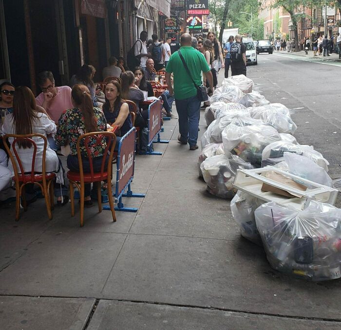 Outdoor diners seated next to lined-up garbage bags on a busy New York street showing city contrasts.