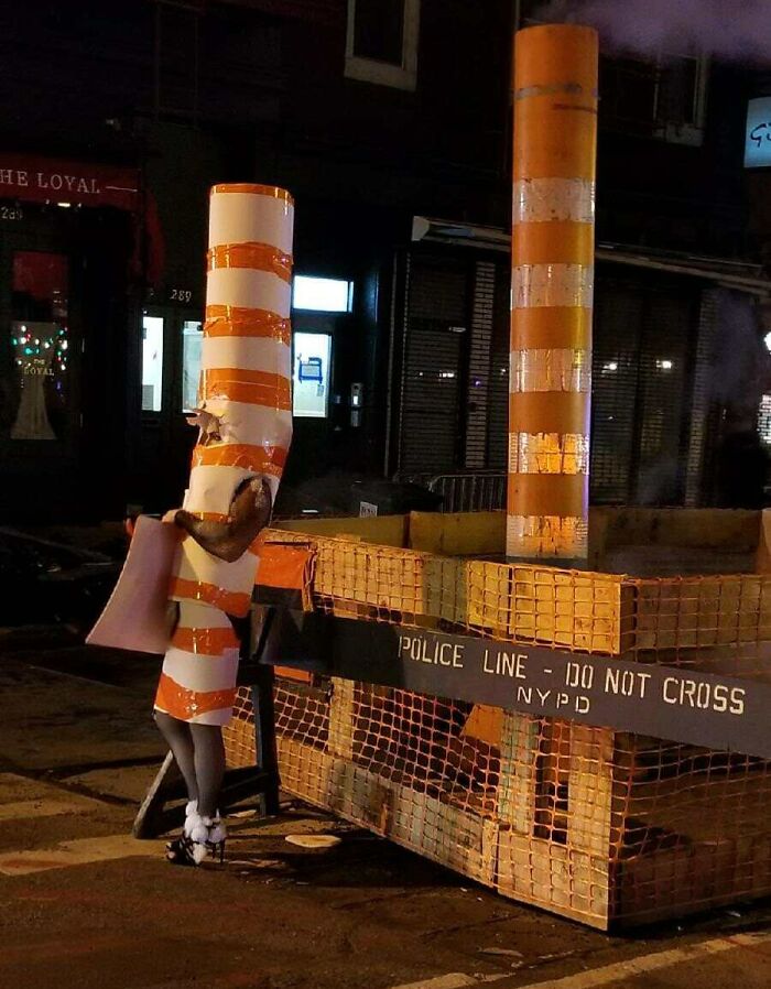 Person dressed as a striped orange and white steam pipe on a New York City street at night near NYPD police line barriers.