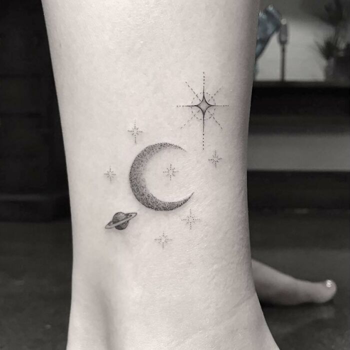 Celebrity tattoo of a crescent moon, stars, and a small planet on an ankle in black ink.