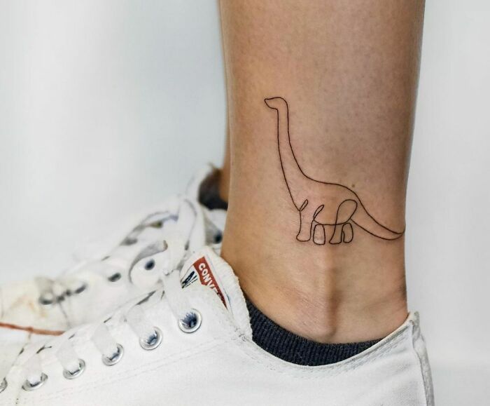 Minimalist dinosaur line tattoo on ankle above white Converse sneaker.