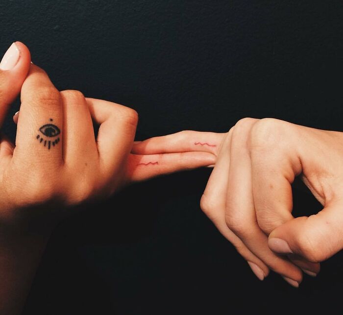 Two hands with celebrity tattoos, one featuring an eye design and the other two matching red waves on fingers.