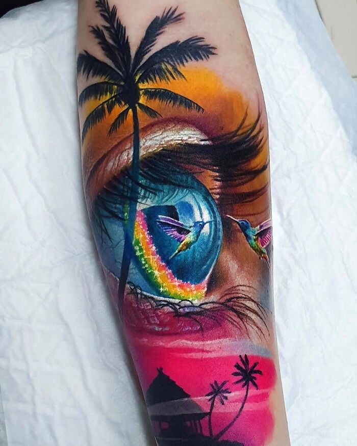 Optical illusion tattoo with eye, palm tree, rainbow, and hummingbirds blending into a colorful design.